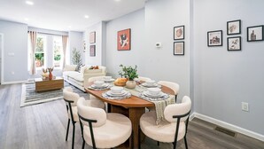 Uma sala de jantar com mesa de madeira posta para quatro pessoas, rodeada de cadeiras, em um ambiente com paredes claras e uma janela grande.