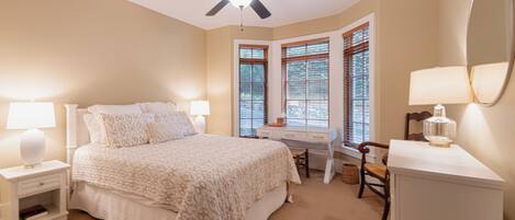 A bedroom with a bed, bedside tables, a desk, a chair, a ceiling fan, and a window with blinds.