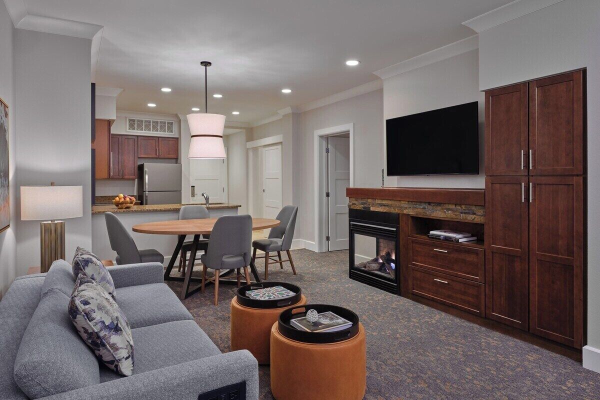 A modern living room with a grey sofa, ottomans, a flat-screen TV, a fireplace, and a dining area with chairs and a table.