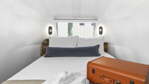 Habitación de hotel pequeña con una cama, almohadas, una maleta y una lámpara fijada a la pared.