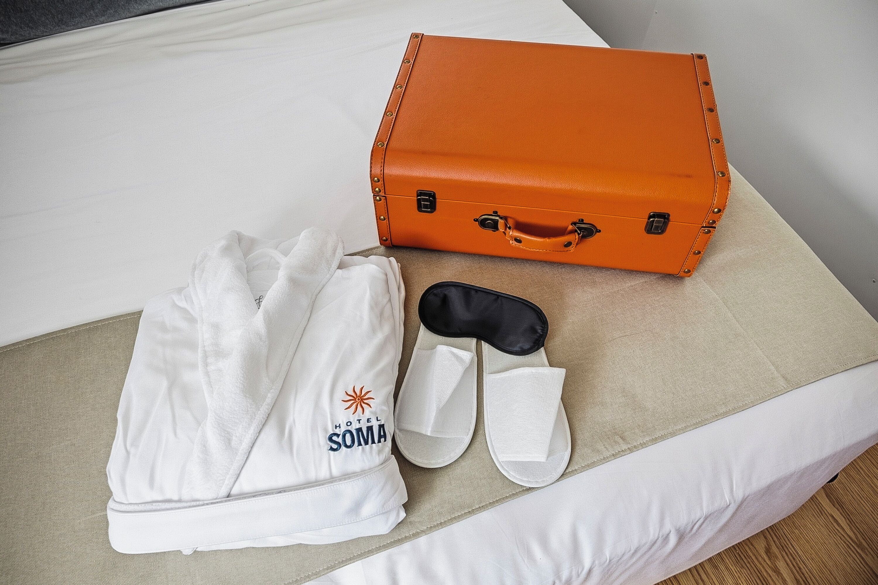 En hvid pude, en hvid morgenkåbe, et par hvide slippers og en orange kuffert på sengen.