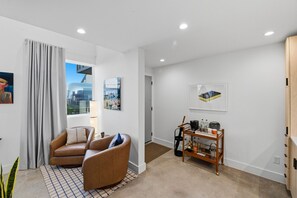 A modern living room with a sofa set, a coffee table, and a side table. There is a window with curtains, a door, and a wall-mounted shelf with various items. - Refined Casita Near Medical District, Parking Included (Fort Worth)