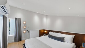 Moderni hotellihuone, jossa on suuri sänky, yöpöytä lampulla, ikkuna verhoilla ja seinään kiinnitetty valaisin.