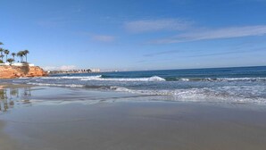 A beach with clear skies, waves approaching the shore, and a distant cityscape. - Casa Kate La Zenia (Orihuela)