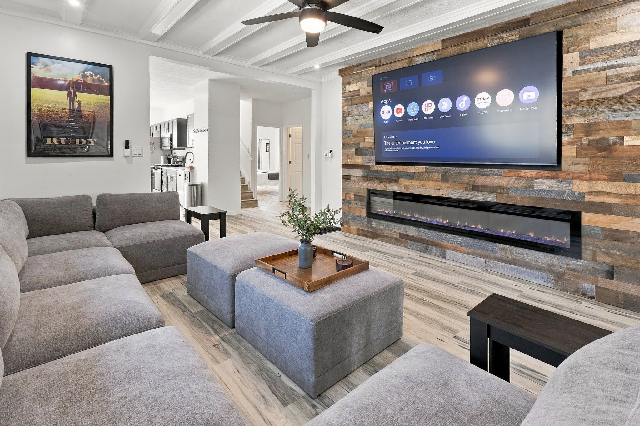 A modern living room with a flat-screen TV mounted on a wooden wall, grey sofas, and a small table with a plant.