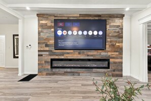A modern living room with a flat-screen TV mounted on a wooden wall, a fireplace below, and a plant in the foreground. - The Social House - 7BR, Hot Tub & Pool Table (Indianapolis)