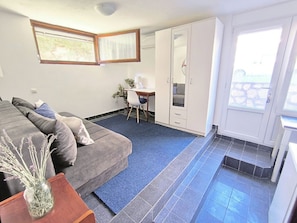 Ein Zimmer mit Sofa, einem kleinen Tisch mit Vase, Schreibtisch mit Stuhl und einem Balkon mit Tür.