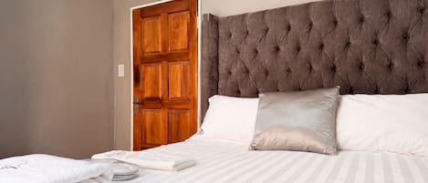 A neatly made bed with striped bedding and two pillows, a wooden door with a clock, and a wall-mounted light switch.