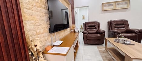 Una sala moderna con una pared de acento de piedra, un televisor de pantalla plana, dos sofás de cuero, una mesa de centro de madera y un jarrón decorativo con flores secas.