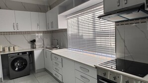 A modern kitchen with a built-in washing machine, a stove, and a window with blinds. - Saob Property-south AF (Germiston)
