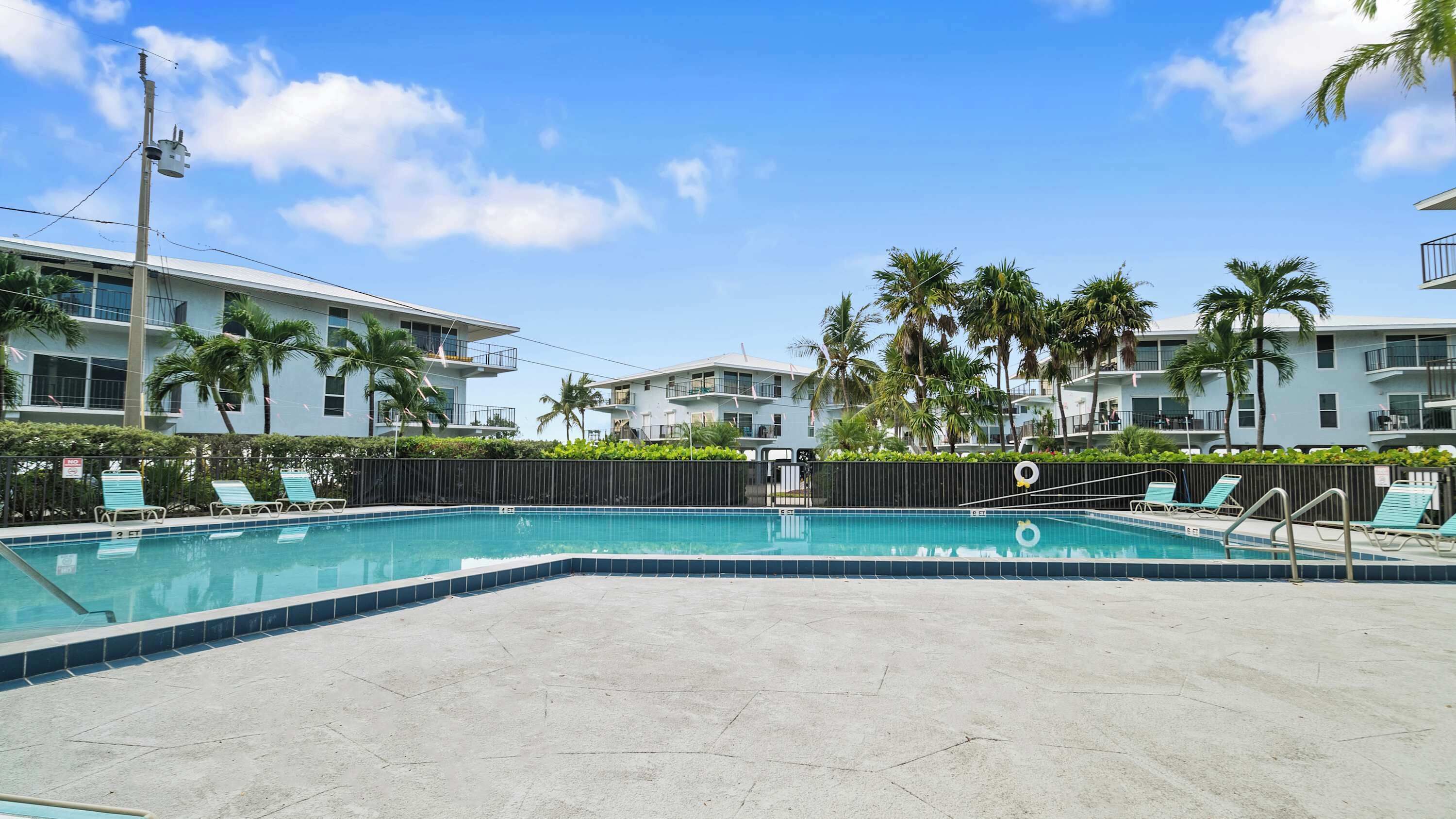 A swimming pool with lounge chairs, surrounded by palm trees and a building with balconies.