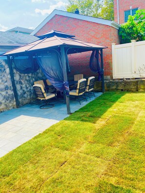 A covered patio area with a stone wall, a white fence, and a grassy lawn. - Queens NYC Private One Bedroom Basement Apartment (Queens)