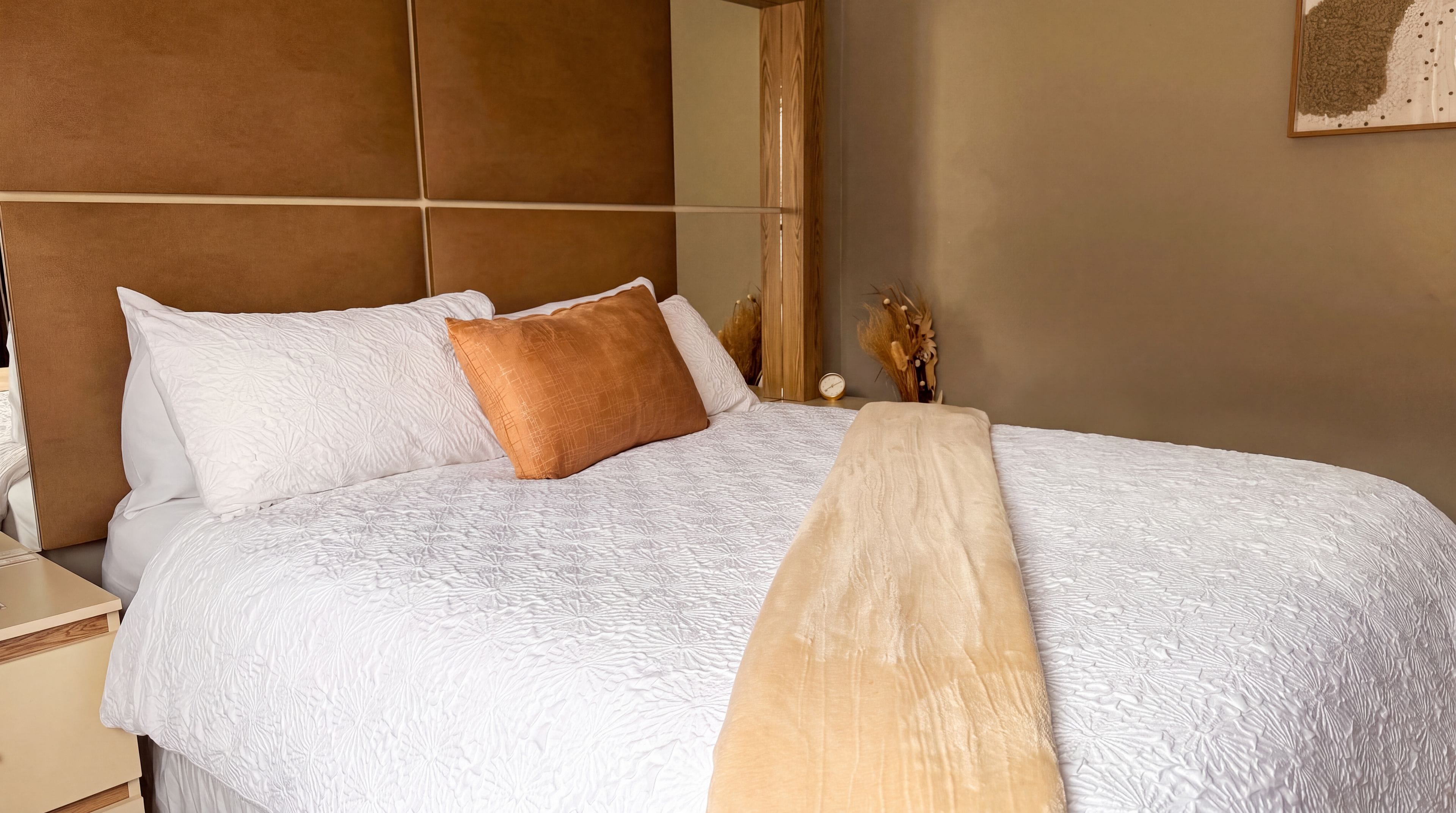 A neatly made bed with a headboard, a bedside table, and a wall clock.