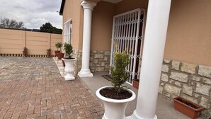 A paved patio with potted plants, a stone wall, and a building with a barred window. - Saob Property South AF (Germiston)