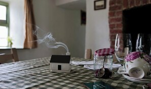 A table with a house-shaped object emitting smoke, a glass jar with a spoon, and a floral teacup on a checkered tablecloth.