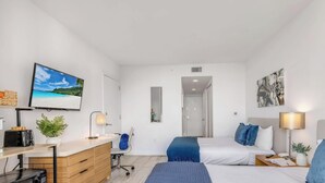 Ein modernes Hotelzimmer mit Bett, Nachttisch, Lampe und Fernseher, der eine Strandansicht zeigt.