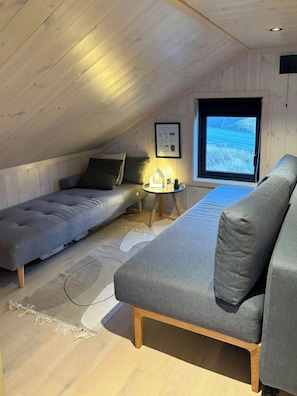 Ein gemütliches Dachzimmer mit Hochbett, Sofa, kleinem Tisch mit Lampe und einem Fenster mit Blick.