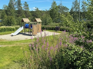 A playground with a slide and climbing structures, surrounded by greenery and flowers.
