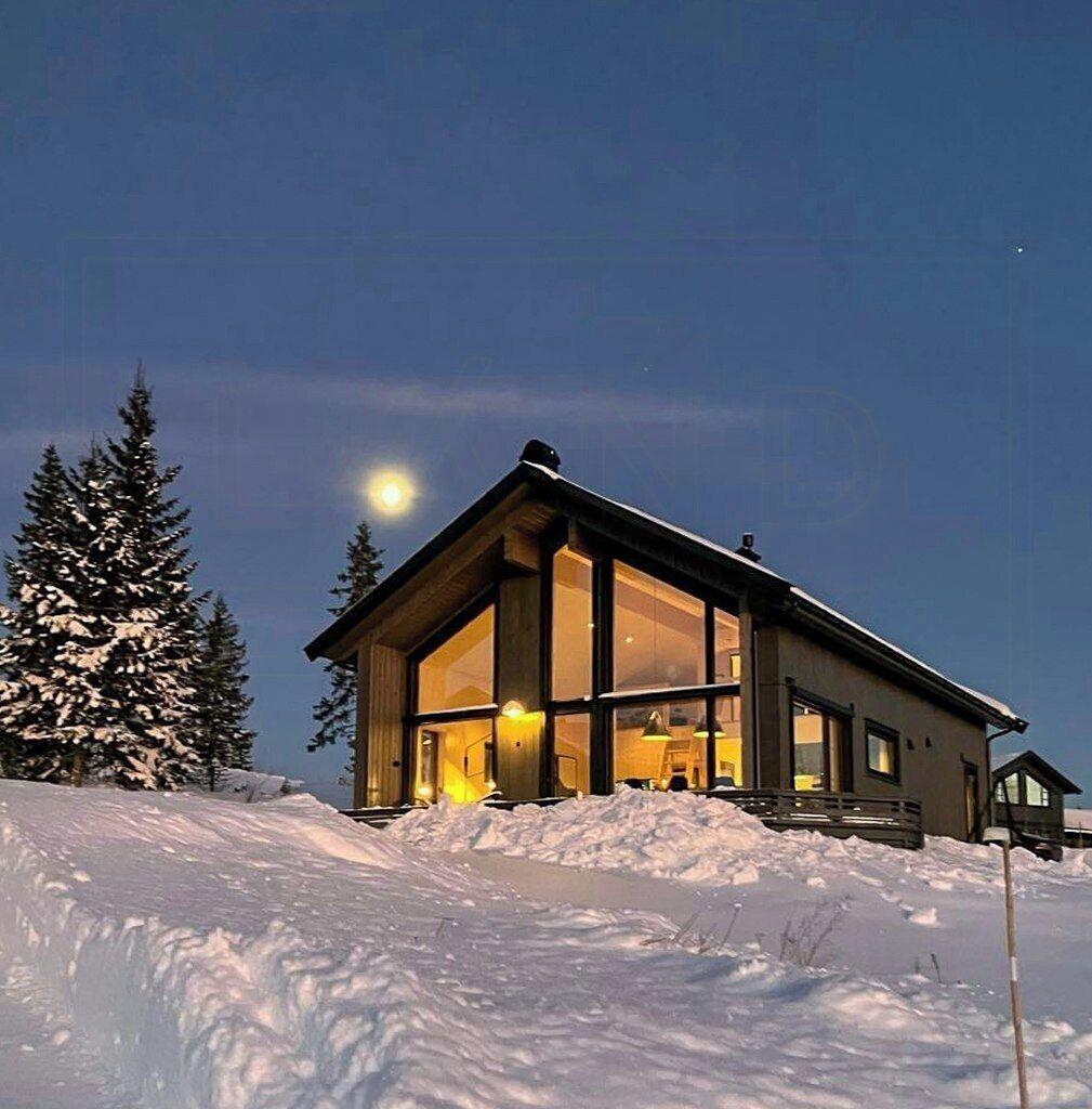 A modern house with large windows, situated on a snow-covered slope with a clear night sky and a visible moon.