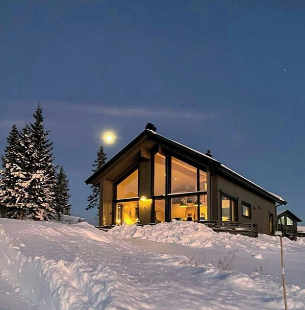 Une maison moderne dotée de grandes fenêtres, située sur une pente enneigée, sous un ciel nocturne dégagé et avec une lune visible.