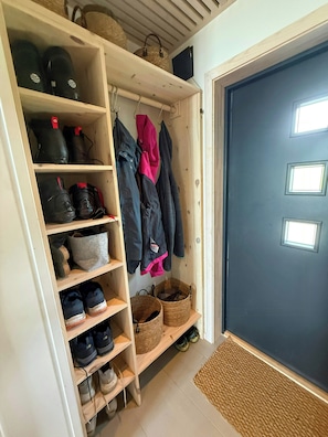 A wooden shoe rack with various footwear, a coat rack with jackets, and a dark door with a window.