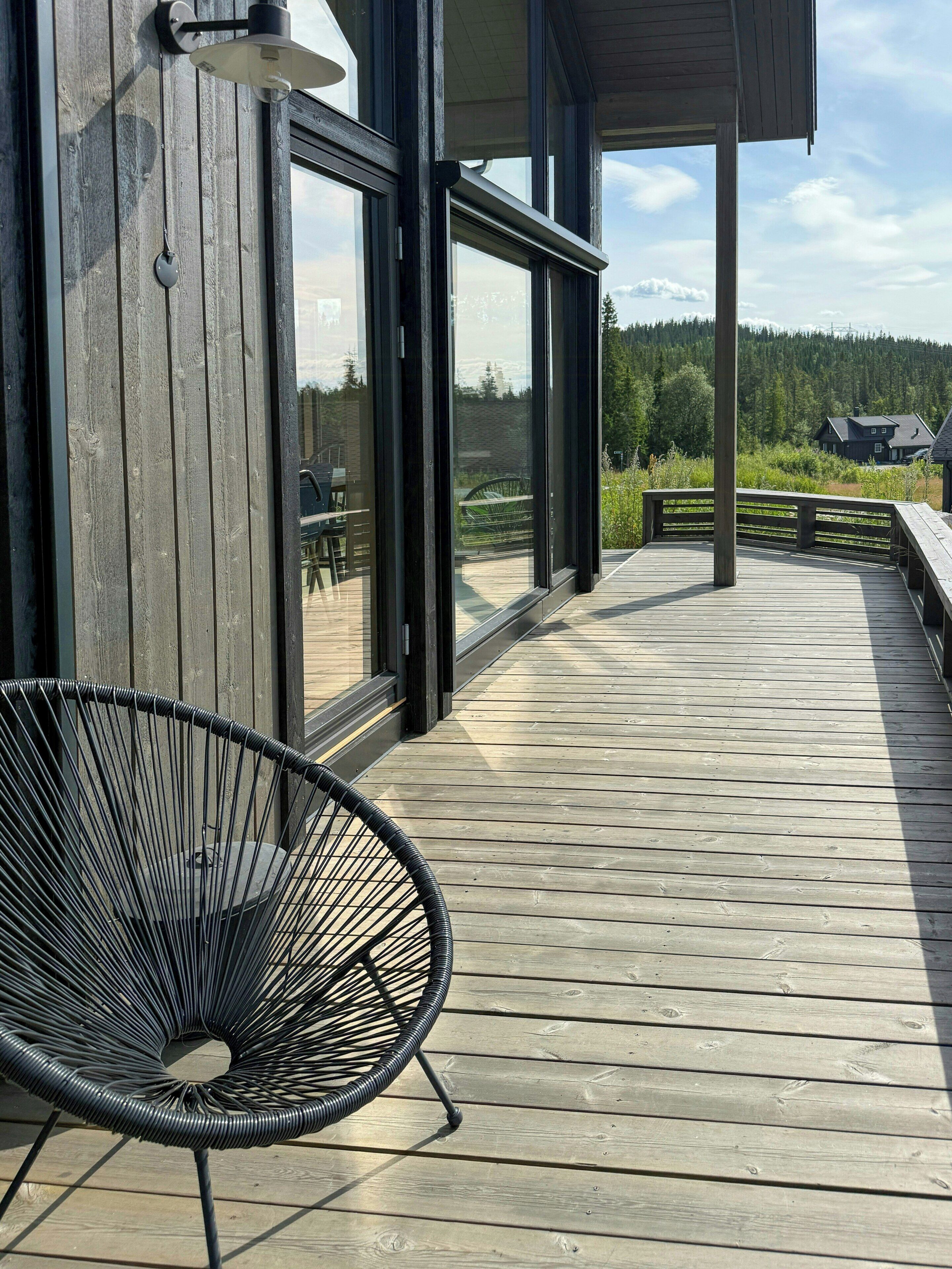 A wooden deck with a black metal chair, large windows, and a view of a forested area.