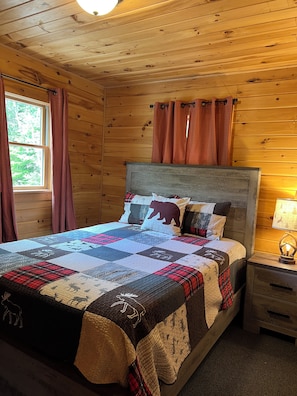 A wooden cabin bedroom with a bed, curtains, a nightstand, and a window.