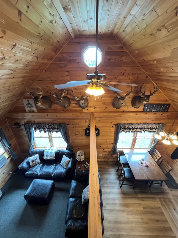 A cabin interior with a high ceiling, wooden walls, a ceiling fan, a dining area, and a sofa with a pillow.