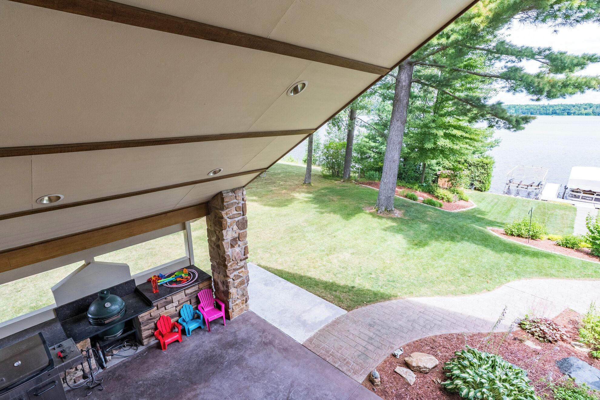 A covered outdoor area with a barbecue grill, colorful Adirondack chairs, and a view of a lake.