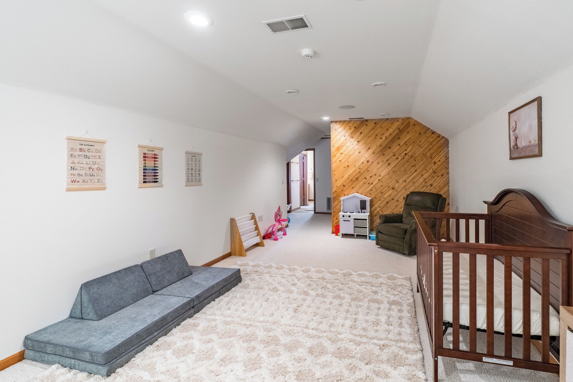 Amplia habitación con un sofá gris, una cuna de madera y una alfombra blanca.