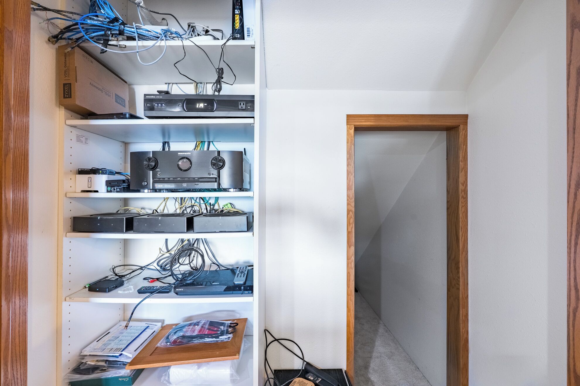 A room with a white wall, a wooden door, and a shelf with electronic devices and cables.