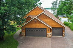 Ein zweistöckiges Holzhaus mit Garage, umgeben von Bäumen und Blick auf einen See.