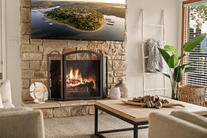 Une cheminée en pierre avec un téléviseur fixé au-dessus, un feu qui crépite à l’intérieur, et une vue sur un lac et une île qui se reflète sur l’écran du téléviseur.