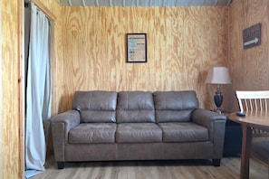 A room with a brown leather sofa, a wooden wall, a framed fish knowledge chart, a table with a lamp, and a directional sign.