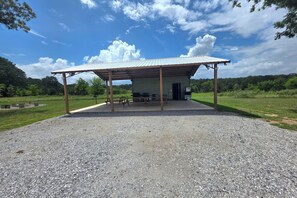 A covered outdoor area with a gravel floor, picnic tables, and a building with a metal roof.