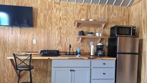 Una cocina compacta con pared de madera, un televisor de pantalla plana, un refrigerador de acero inoxidable, una cafetera y un taburete de bar.