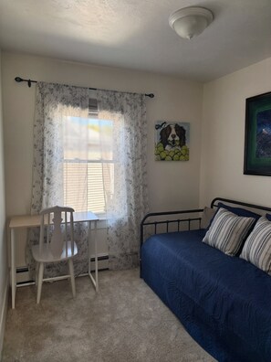 A bedroom with a bed, a desk, a chair, a window with curtains, and a painting on the wall.