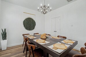 Ein Esszimmer mit einem langen Tisch, gedeckt für sechs Personen, umgeben von Holzstühlen, einer Kristallleuchtre und einem Wandbehang.