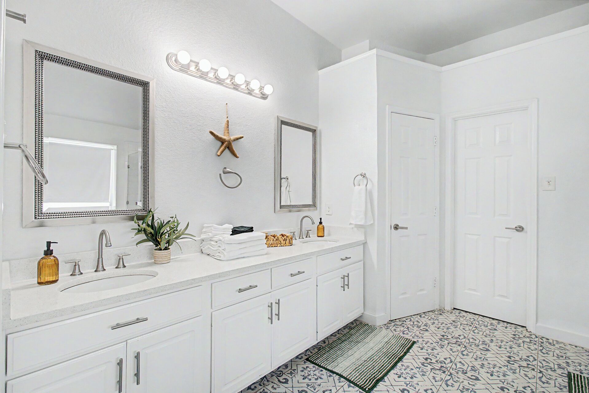 Une salle de bain avec un meuble-lavabo blanc, deux miroirs, une décoration en forme d’étoile de mer, un porte-serviettes et un tapis à motifs.
