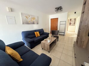 Un salon moderne avec un canapé bleu, une table basse en bois et un fauteuil gris. Une carte du monde est accrochée au mur, et une porte donne accès à une autre pièce.