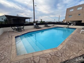 Ein rechteckiger Pool mit einer gefliesten Kante, umgeben von einer Terrasse mit Steinmuster. Auf der Terrasse stehen Liegestühle und ein kleiner Tisch.