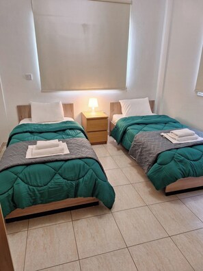 Ein Doppelzimmer mit zwei Betten, jedes mit einer grünen Bettdecke, einem Nachttisch mit Lampe und einem Fenster über den Betten.