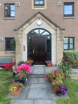 Ein Gebäude mit einer Stein-Fassade, einer dunkelbraunen Eingangstür und der Hausnummer 63 darüber. Vor dem Eingang stehen Pflanzgefäße mit verschiedenen Blumen.
