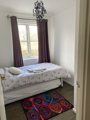 Ein Schlafzimmer mit einem Bett, einem Fenster mit Vorhängen, einer Kristallleuchtre und einem farbenfrohen Teppich.