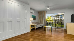 Quarto com piso de madeira, sofá, mesa pequena, luminária, TV e varanda com vista para prédios e palmeiras.