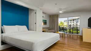 Ett hotellrum med en stor säng, en soffa, ett litet bord och en balkong med utsikt över en byggnad och grönområden.