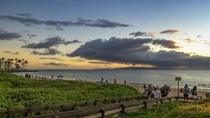 Uma praia com um deck de madeira, pessoas caminhando e uma placa com a inscrição 'CUIDADO'. 