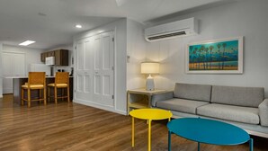 Uma sala de estar moderna com um sofá cinzento, uma mesa de centro azul e uma mesa lateral amarela. Há um quadro com palmeiras na parede, e um aparelho de ar condicionado montado acima do sofá.