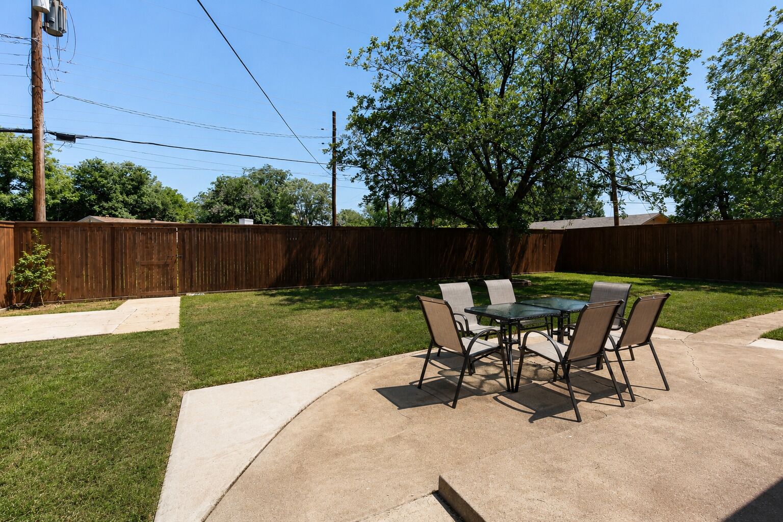 A backyard patio with a table and chairs, surrounded by a wooden fence and a tree.