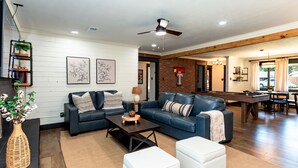A modern living room with a sofa, coffee table, and ottomans, a dining area with a pool table, and a kitchen visible through an open doorway.